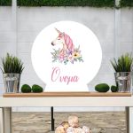 Ξύλινη στρογγυλή πινακίδα, Μονόκερος ακουαρέλα με ζωγραφιστά λουλούδια, με προσαρμοσμένο όνομα, σε τραπέζι.