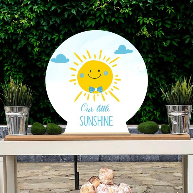 Wooden Baptism/candybar figure: happy sun boy with blue bow tie, "Our little Sunshine". White table, green backdrop.