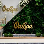 Gold metal baptism arch with hanging name 'Faidra' in gold plexiglass, against a plant background.