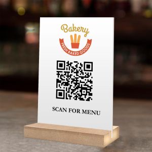 White plexiglass QR menu sign with a Bakery logo, standing on a wooden base.