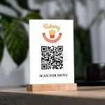 White plexiglass QR menu sign with a logo, set in a light wooden base on a tabletop.