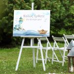 Baptism welcome canvas featuring lighthouse, paper boat, waves. On white easel, set outdoors on grass.