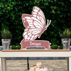 Minimalist wooden butterfly, muted rose with name, standing on wooden shelf against lush green ivy wall.