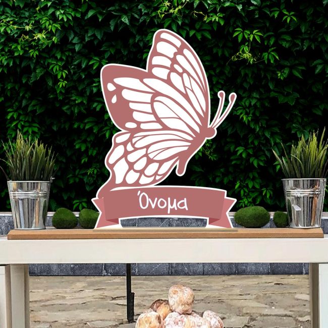 Minimalist wooden butterfly, muted rose with name, standing on wooden shelf against lush green ivy wall.