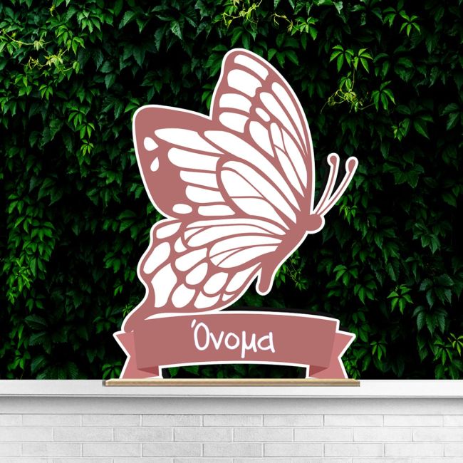 Minimalist wooden butterfly, dusky pink, personalized, on a wooden shelf against a green plant wall and white brick.