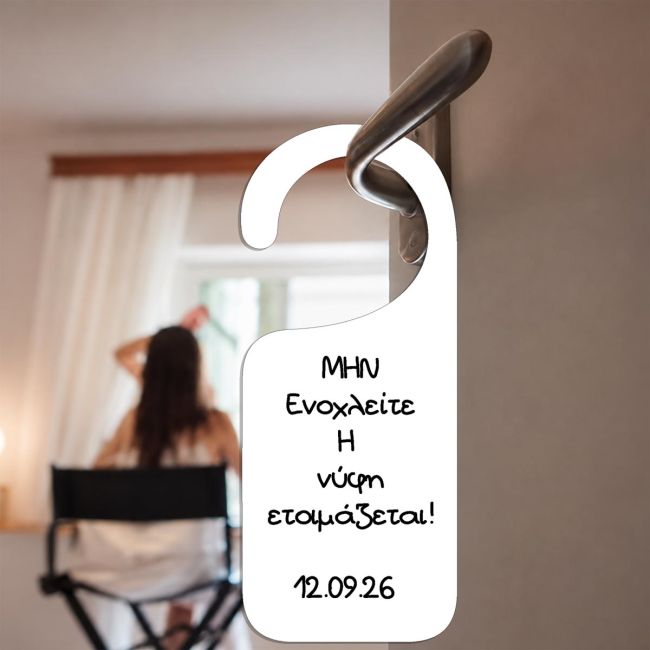 Λευκή κρεμάστρα πόρτας 'Η νύφη ετοιμάζεται', εκτυπωμένη σε πλαστικό/χαρτόνι, κρεμασμένη σε πόμολο, με νύφη στο βάθος.