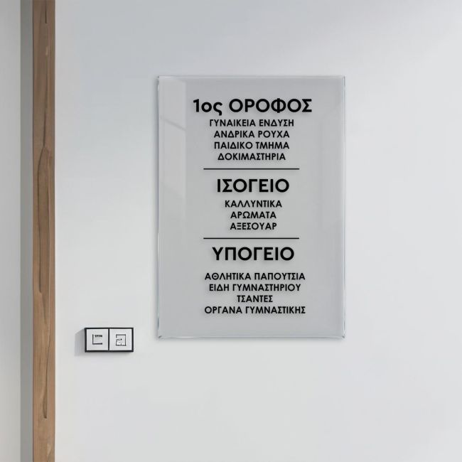 Διάφανη ακρυλική πινακίδα ορόφων καταστήματος σε λευκό τοίχο με μαύρα γράμματα.